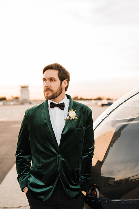 Groom in an ivory paisley tuxedo gently holds his bride’s face as they smile at each other on the airstrip.
