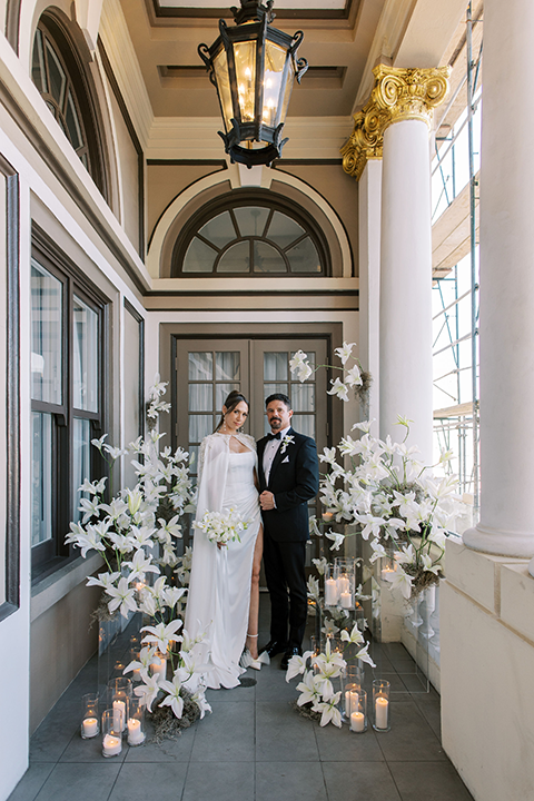 A newlywed couple poses in a romantic white and ivory wedding setting, surrounded by elegant white lilies, glowing candles, and sheer drapery.