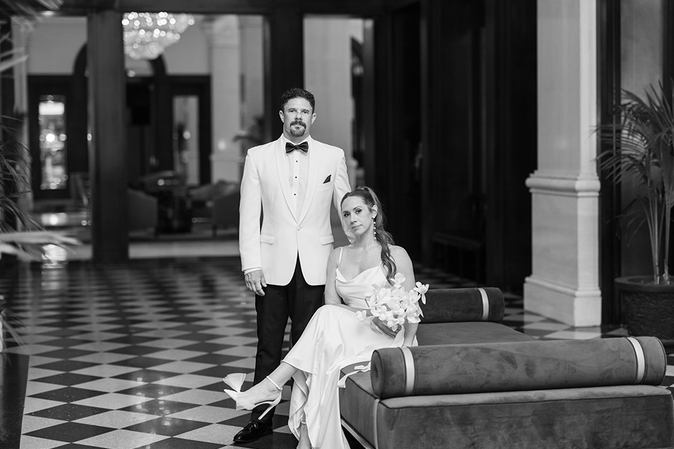 A newlywed couple poses in a romantic white and ivory wedding setting, surrounded by elegant white lilies, glowing candles, and sheer drapery. The bride wears a lace gown with a cape, while the groom wears a white tuxedo jacket with black trousers.