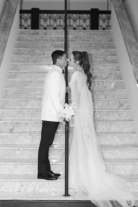 A newlywed couple poses in a romantic white and ivory wedding setting, surrounded by elegant white lilies, glowing candles, and sheer drapery. The bride wears a lace gown with a cape, while the groom wears a white tuxedo jacket with black trousers.