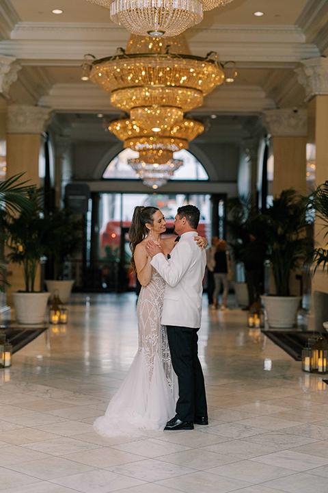 A newlywed couple poses in a romantic white and ivory wedding setting, surrounded by elegant white lilies, glowing candles, and sheer drapery. The bride wears a lace gown with a cape, while the groom wears a white tuxedo jacket with black trousers.