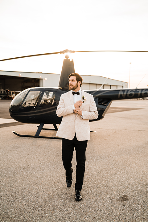 Groom in an ivory paisley tuxedo gently holds his bride’s face as they smile at each other on the airstrip.