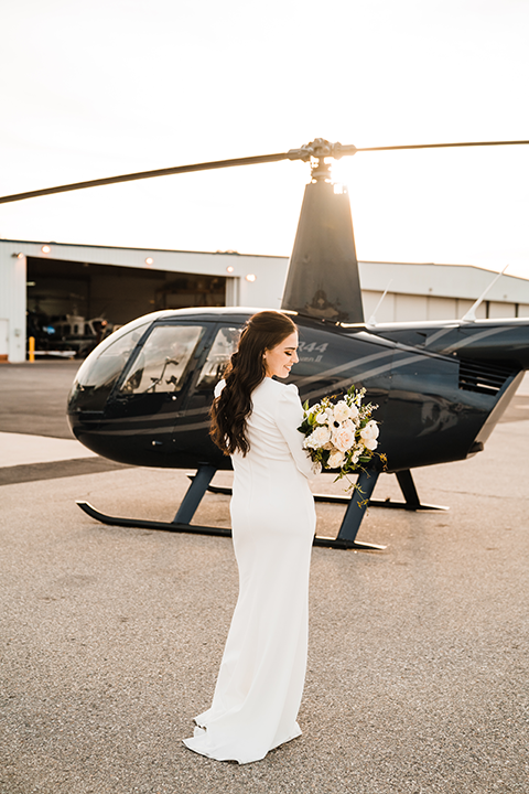 Bride smiles softly, her long-sleeve gown and chic bouquet complementing the aviation-inspired setting