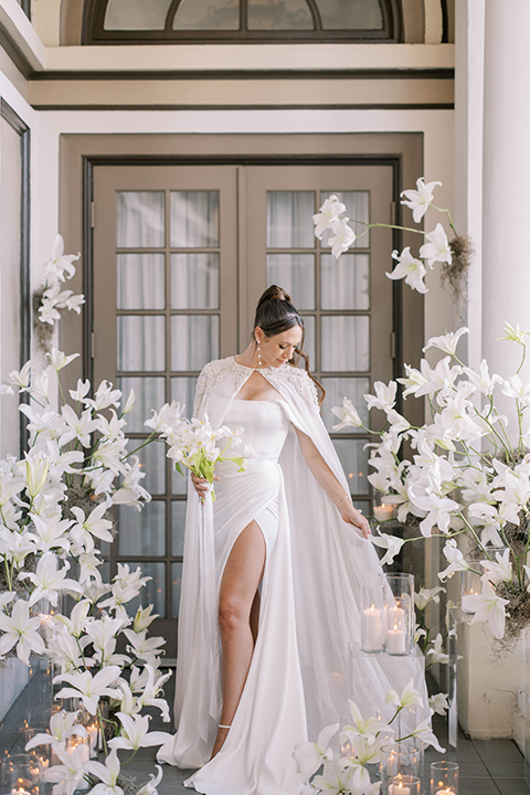 The bride stands elegantly at the historic US Grant House, wearing a sheer lace gown with delicate embellishments and a flowing cape. 
