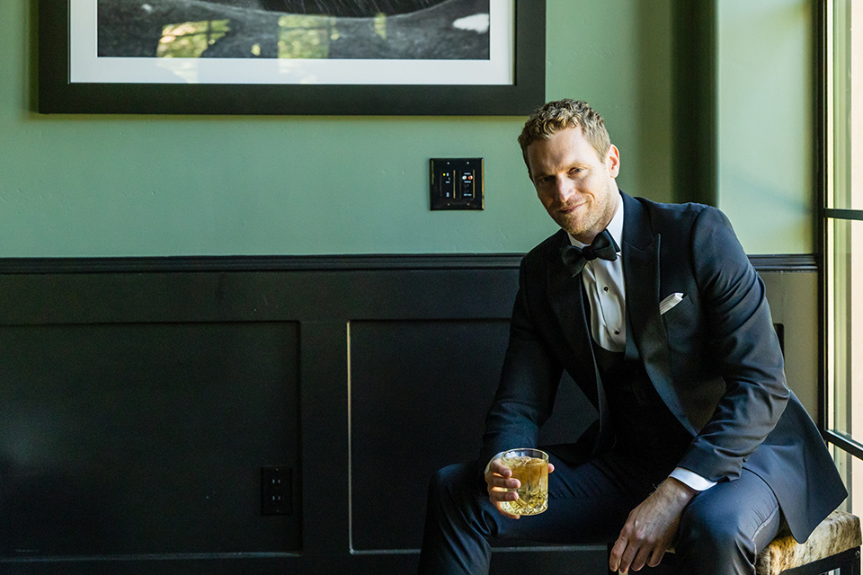 groom in black tuxedo sitting looking at the camera with a glass of whiskey in his hand