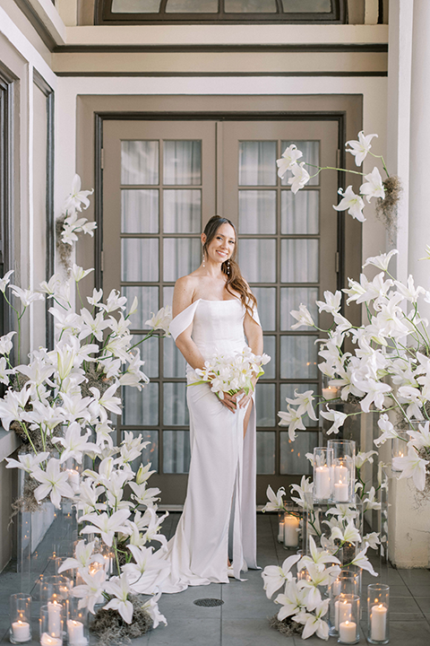 The bride stands elegantly at the historic US Grant House, wearing a sheer lace gown with delicate embellishments and a flowing cape. She holds a bouquet of crisp white florals, exuding timeless beauty and grace.