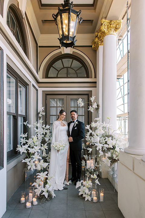 A newlywed couple poses in a romantic white and ivory wedding setting, surrounded by elegant white lilies, glowing candles, and sheer drapery.