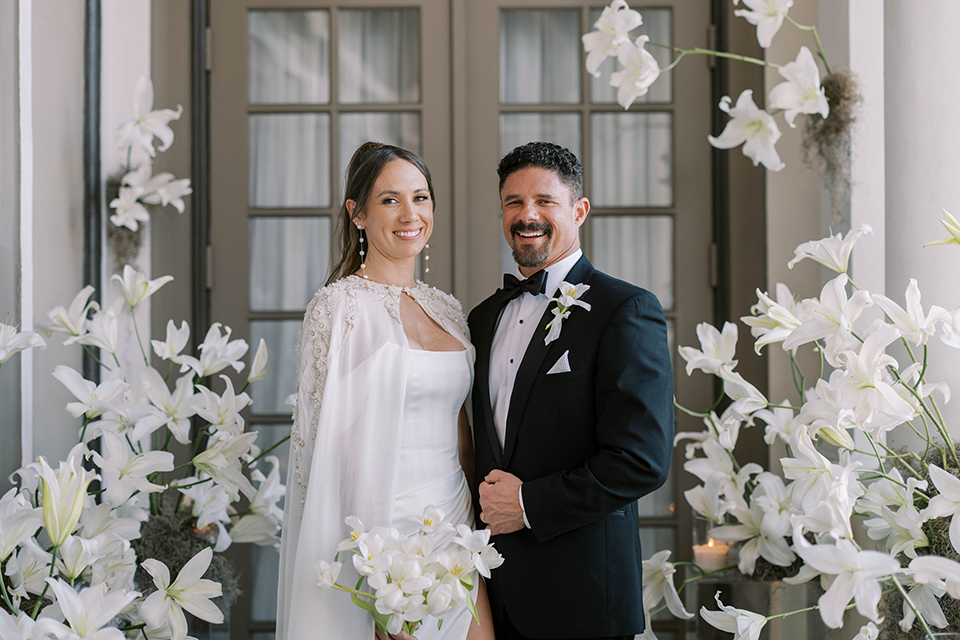 A newlywed couple poses in a romantic white and ivory wedding setting, surrounded by elegant white lilies, glowing candles, and sheer drapery.